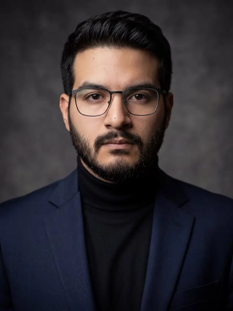 Portrait of Jeremy Orellana: a bearded man with glasses, in a dark blue blazer and black turtleneck, against a dark grey studio background.